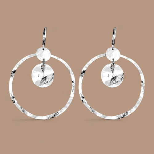 Grandes boucles d'oreilles créoles argentées avec sequins en métal martelé, avec attaches-dormeuses, sur fond beige