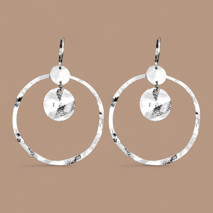 Grandes boucles d'oreilles créoles argentées avec sequins en métal martelé, avec attaches-dormeuses, sur fond beige