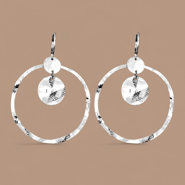 Grandes boucles d'oreilles créoles argentées avec sequins en métal martelé, avec attaches-dormeuses, sur fond beige