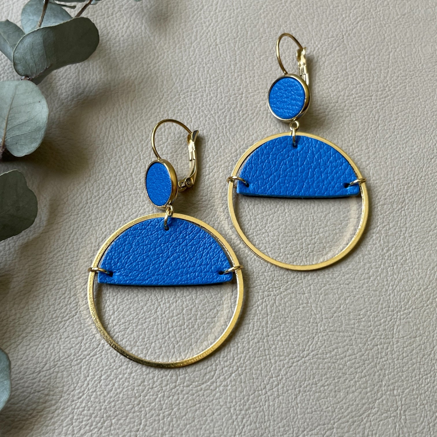 Paire de boucles d'oreilles créoles dorées Nune modèle Lua avec cuir bleu roi, sur fond cuir beige doré