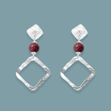 Charger l&#39;image dans la galerie, Boucles d&#39;oreilles losanges sur puces en métal martelé argenté et perles de Mokaïte bordeaux, sur fond bleu gris
