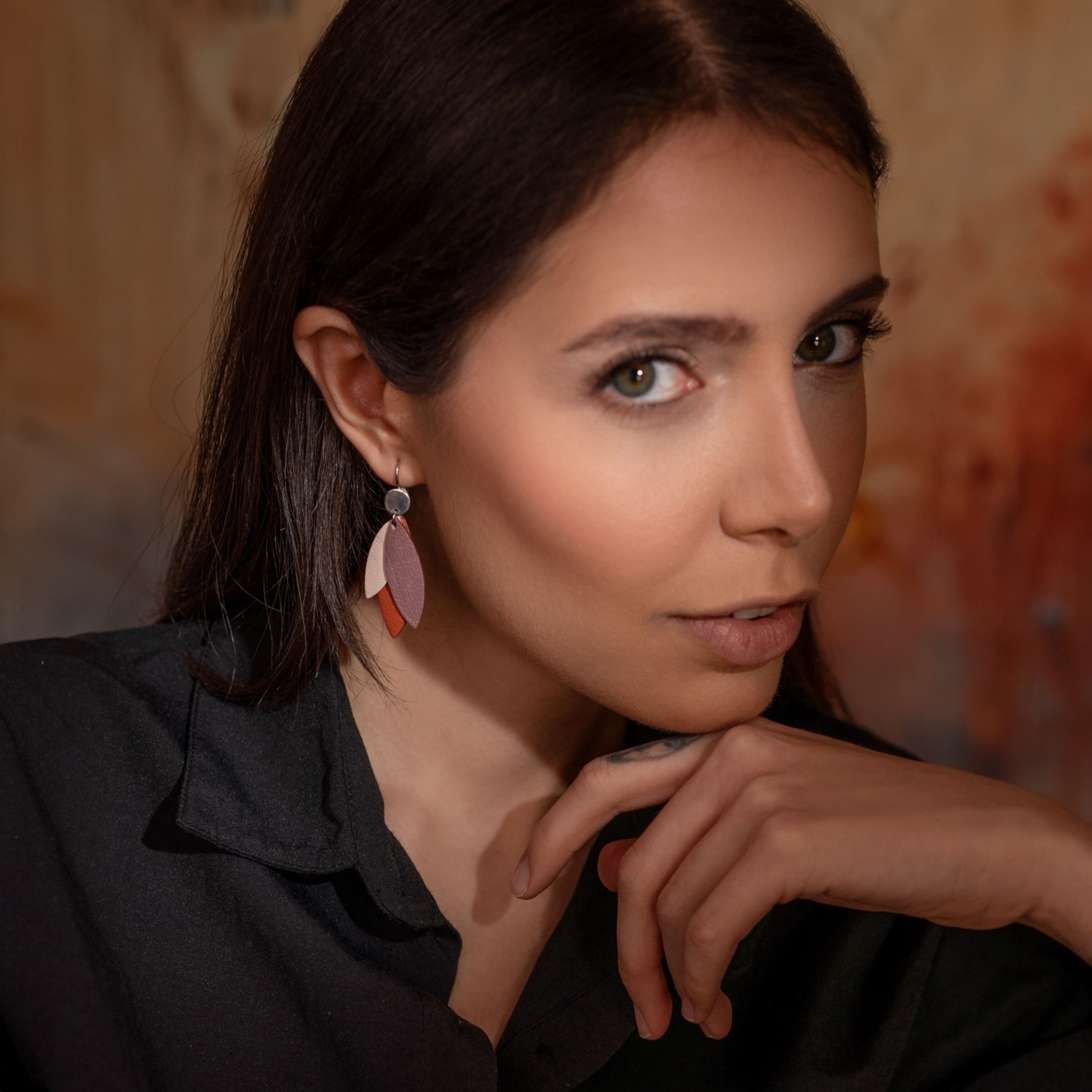 Boucles d'oreilles pendantes argentées et plumes de cuir dans les tons rosés, portées à l'oreille par une femme brune