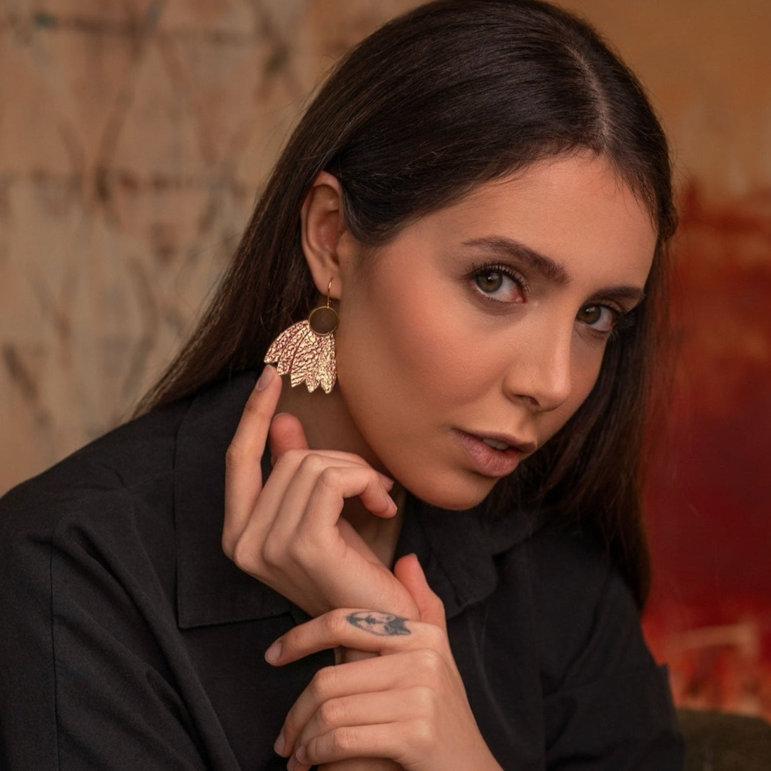 Grandes boucles d'oreilles dorées en forme de couronne de plumes en cuir or rosé froissé,  portées à l'oreille par une femme brune