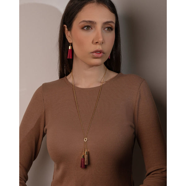 Femme brune portant des boucles d'oreilles dorées en pompons de cuir framboise et portant un collier- sautoir doré avec 2 pompons en cuirs pourpre et fauve or façon serpent