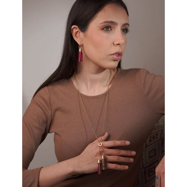 Femme brune portant des boucles d'oreilles dorées en pompons de cuir framboise et soulevant un collier- sautoir doré avec 2 pompons en cuirs pourpre et fauve or façon serpent