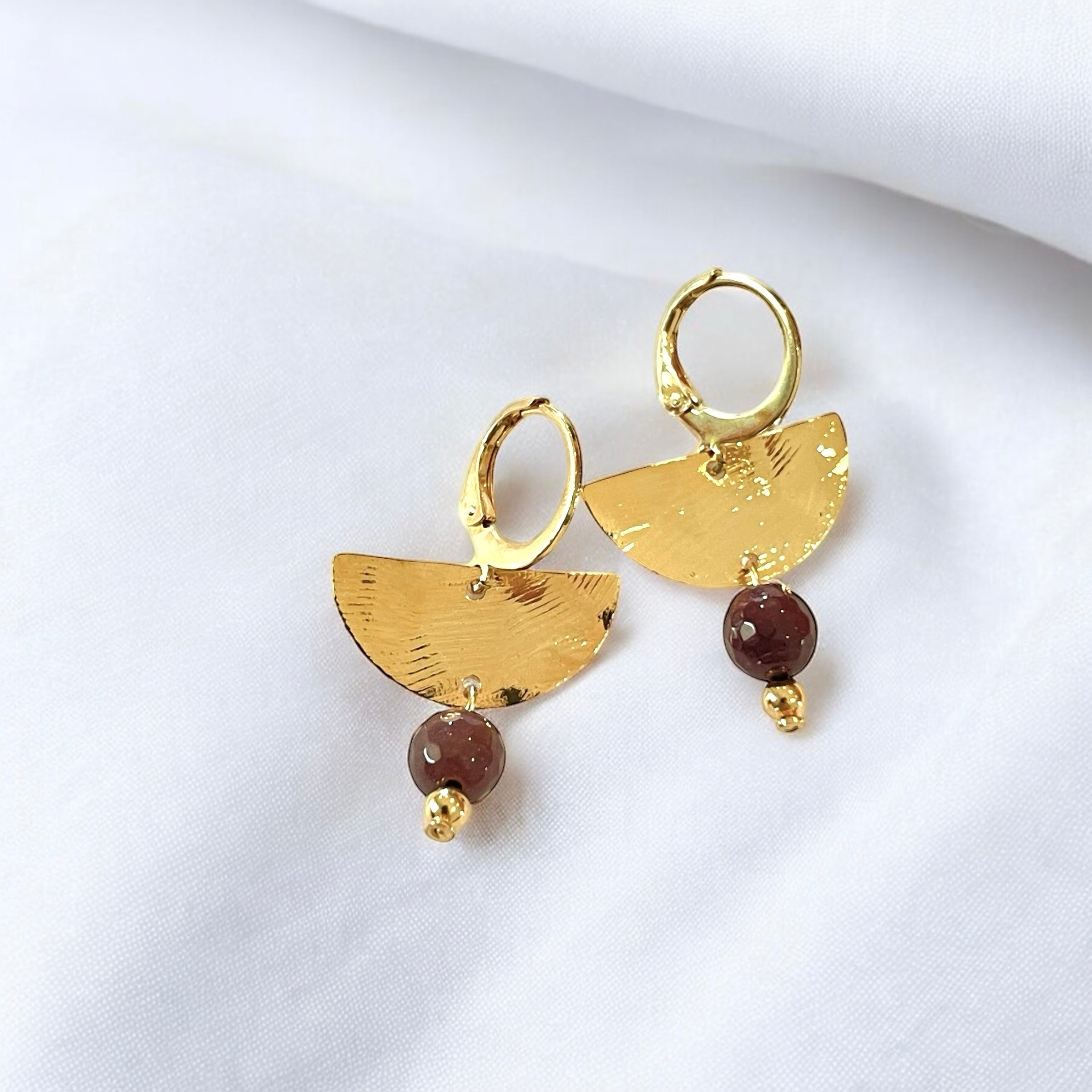 Petites boucles d'oreilles pendantes, de forme demi-lune, en métal martelé doré à l'or fin et perles de Mokaïte, sur un tissublanc