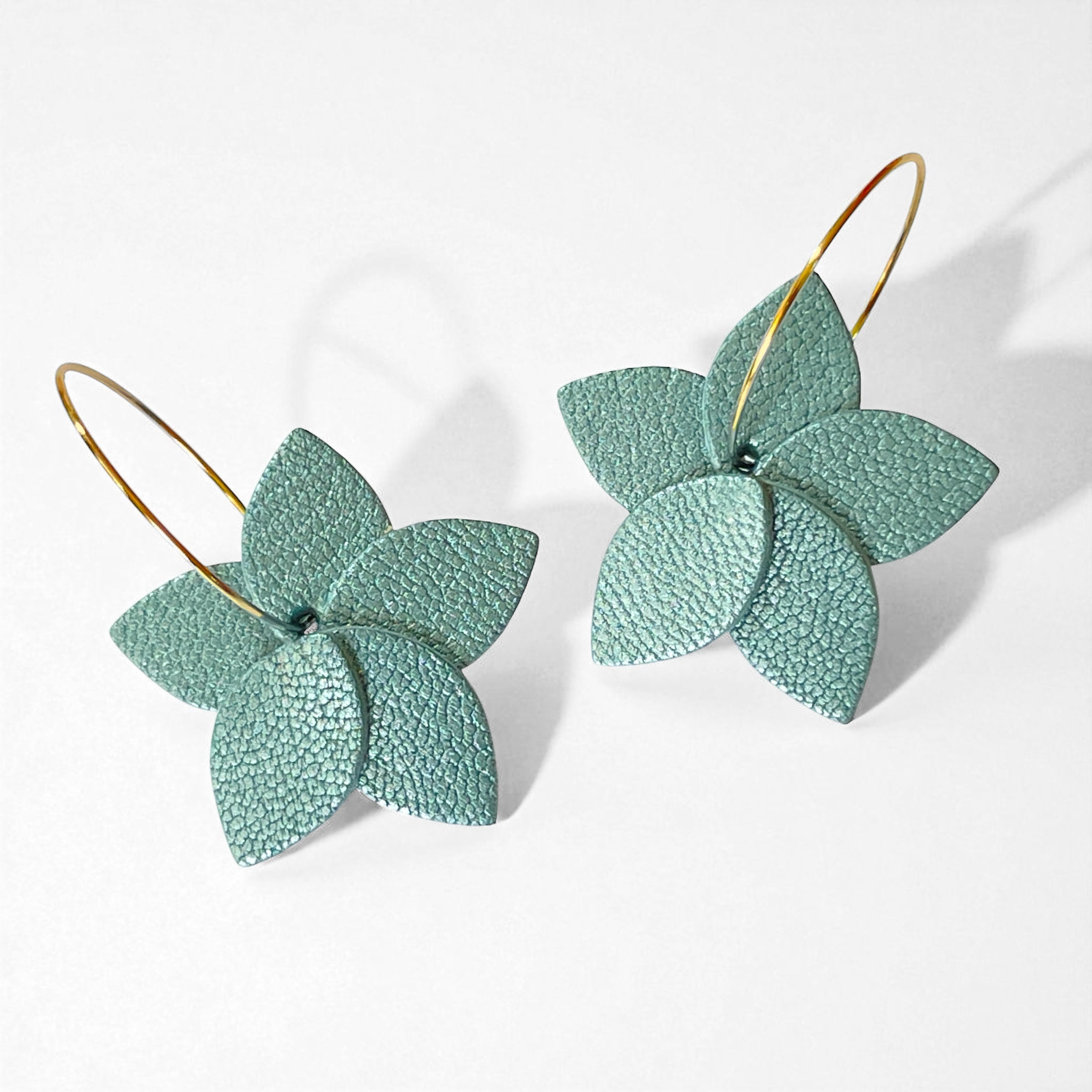 Petites boucles d'oreilles en forme de fleurs en cuir bleu gris nacré, montées sur créoles dorées, sur fond blanc