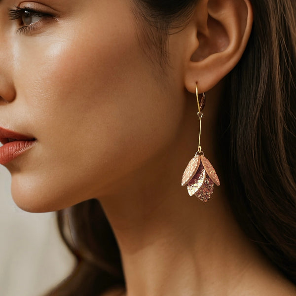 Boucles d'oreilles dorées longues et fines terminées par une grappe de pétales de cuir or rosé froissé, portées à l'oreille par une femme brune
