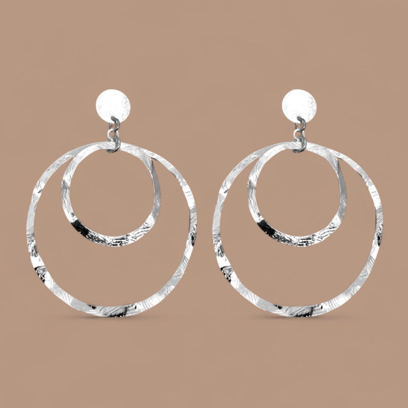 Grandes boucles d'oreilles créoles argentées à double anneau en métal martelé, montées sur puces, sur fond beige