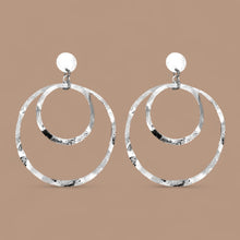 Charger l&#39;image dans la galerie, Grandes boucles d&#39;oreilles créoles argentées à double anneau en métal martelé, montées sur puces, sur fond beige
