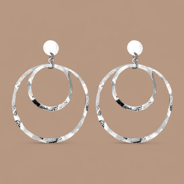 Grandes boucles d'oreilles créoles argentées à double anneau en métal martelé, montées sur puces, sur fond beige