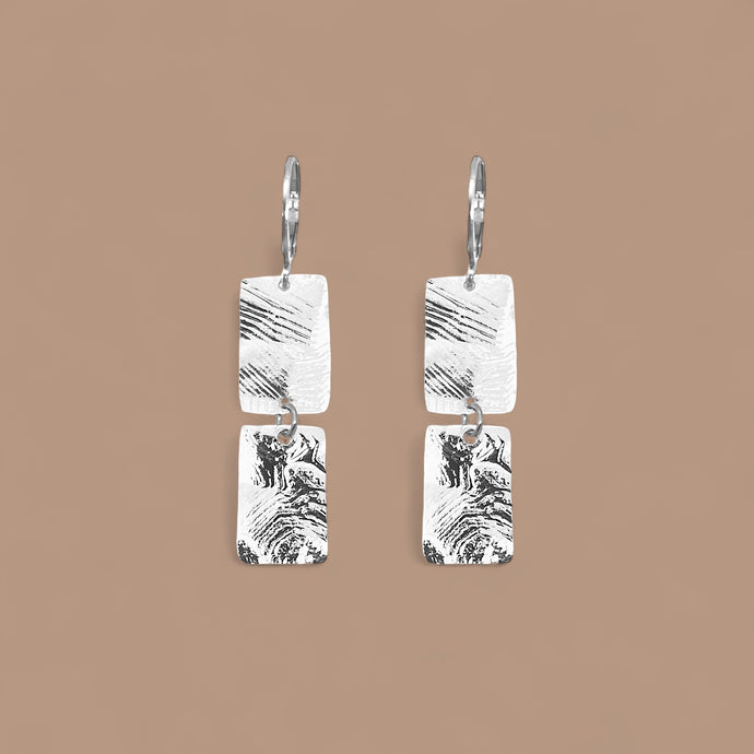 Boucles d'oreilles pendantes composées de 2 rectangles en métal martelé argentées, sur fond beige
