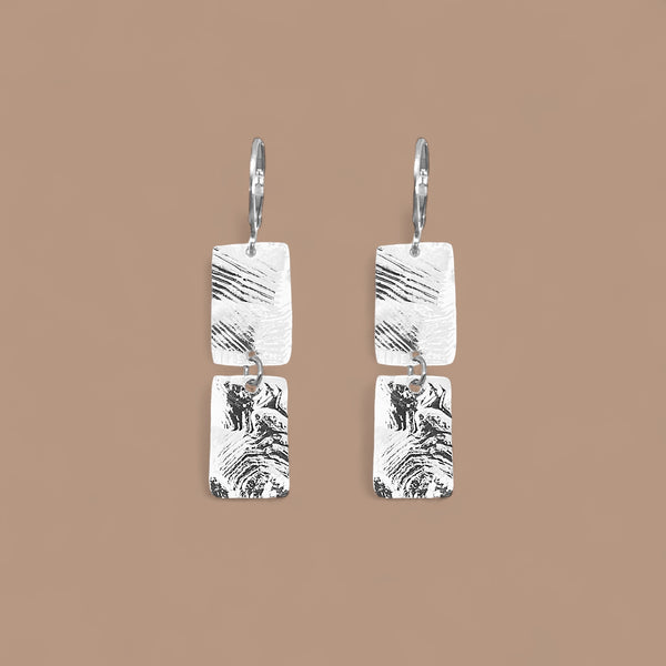 Boucles d'oreilles pendantes composées de 2 rectangles en métal martelé argentées, sur fond beige