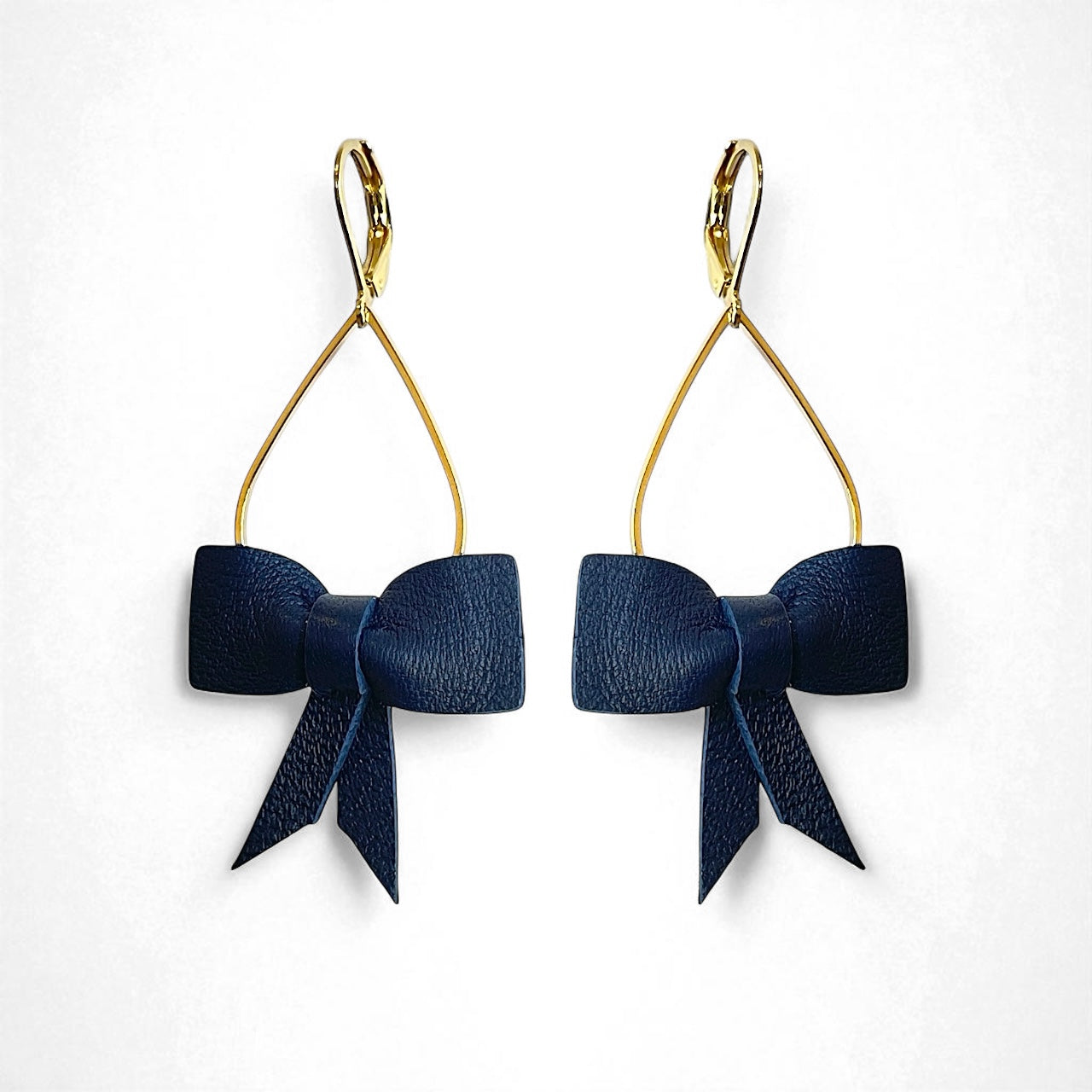 Boucles d'oreilles dorées pendantes et légères en forme de gouttes, décorées de délicats nœuds de rubans de cuir bleu marine satiné, sur fond blanc. Modèle Séraphine de Nune.