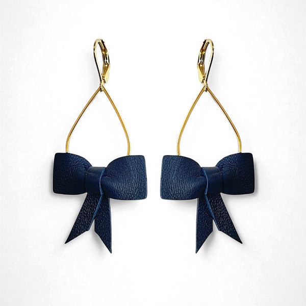 Boucles d'oreilles dorées pendantes et légères en forme de gouttes, décorées de délicats nœuds de rubans de cuir bleu marine satiné, sur fond blanc. Modèle Séraphine de Nune.