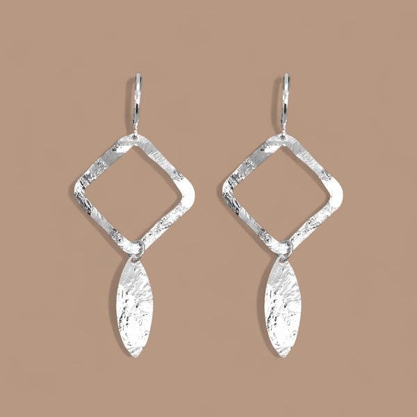 Boucles d'oreilles argentées avec pendentif en forme de losanges et pointes, sur fond beige