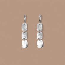 Charger l&#39;image dans la galerie, Fines boucles d&#39;oreilles argentées pendantes en chapelet de 3 petits rectangles, sur fond beige
