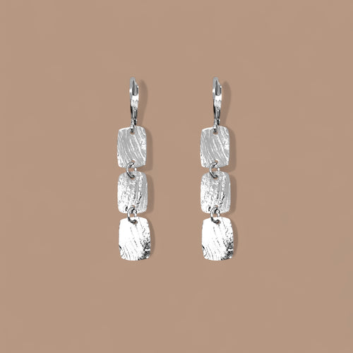 Fines boucles d'oreilles argentées pendantes en chapelet de 3 petits rectangles, sur fond beige