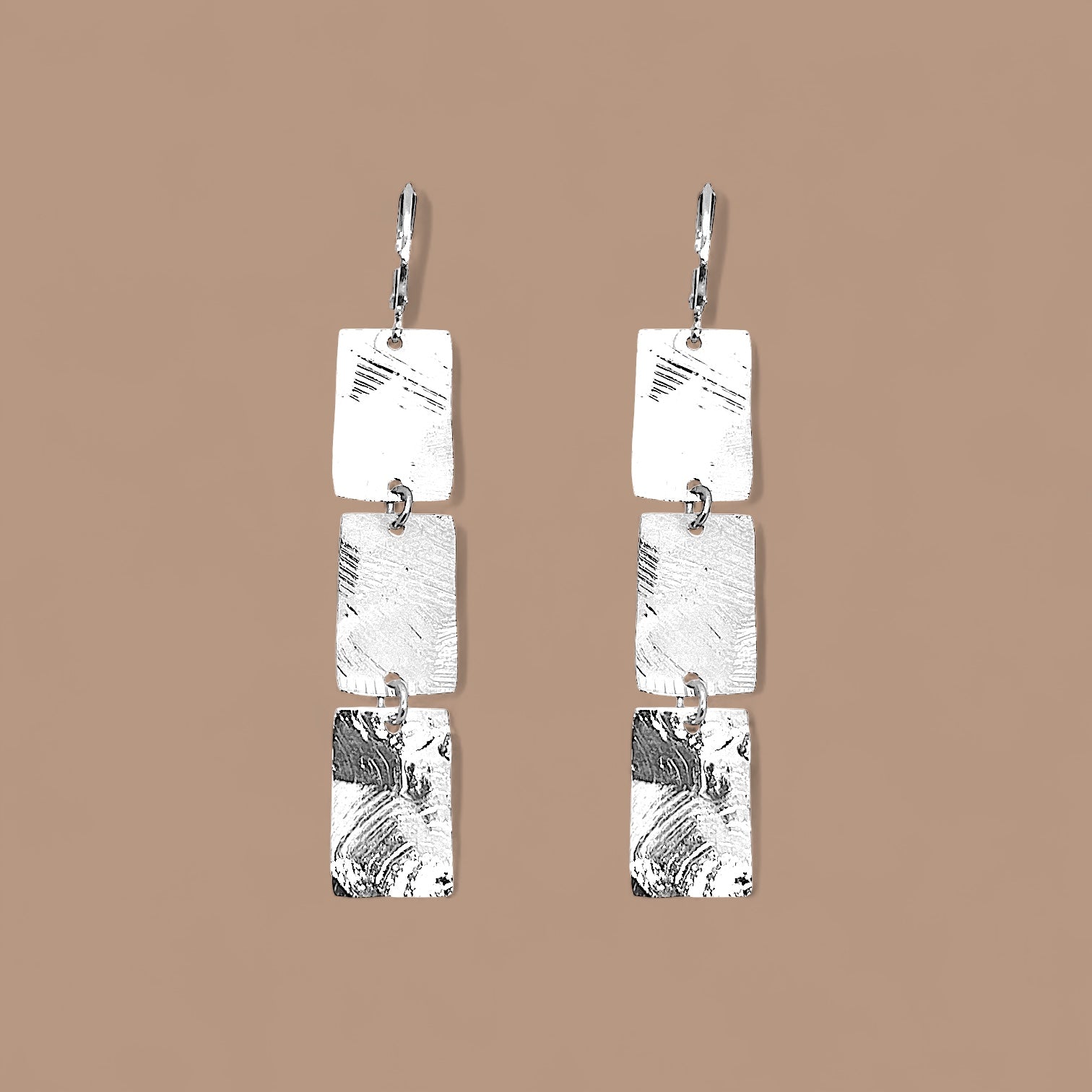 Boucles d'oreilles pendantes en chapelet de 3 rectangles en métal martelé argenté, sur fond beige
