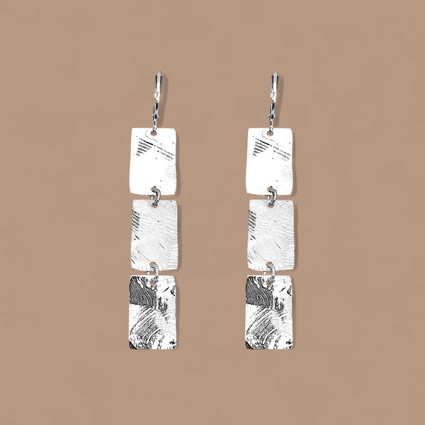 Boucles d'oreilles pendantes en chapelet de 3 rectangles en métal martelé argenté, sur fond beige