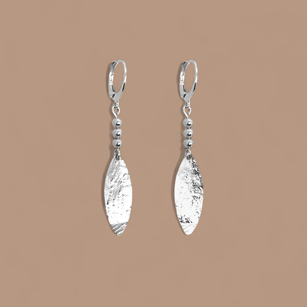 Boucles d'oreilles argentées fines pendantes longues en pointes, sur fond beige