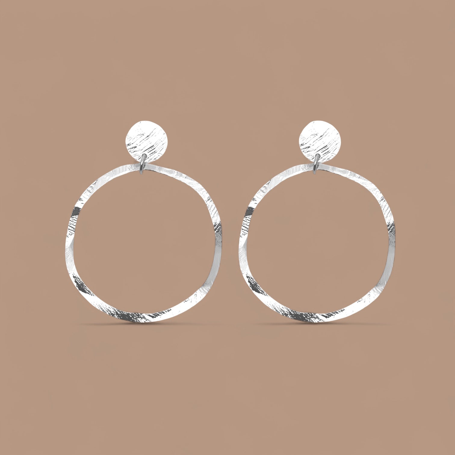 Grandes boucles d'oreilles créoles argentées en métal martelé, montées sur puces rondes, sur fond beige