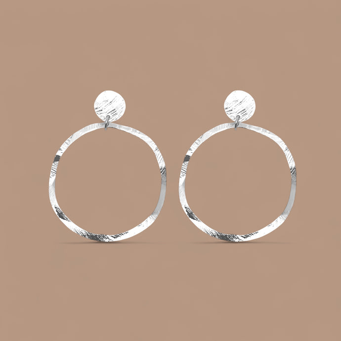 Grandes boucles d'oreilles créoles argentées en métal martelé, montées sur puces rondes, sur fond beige