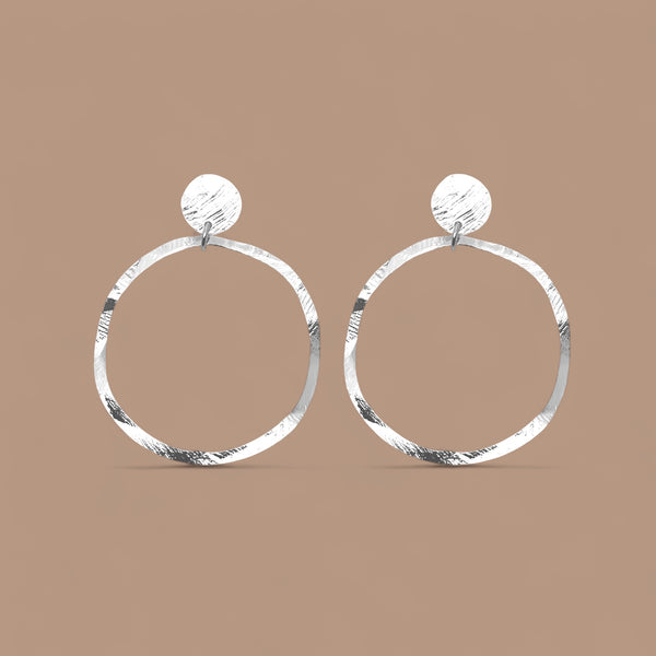 Grandes boucles d'oreilles créoles argentées en métal martelé, montées sur puces rondes, sur fond beige