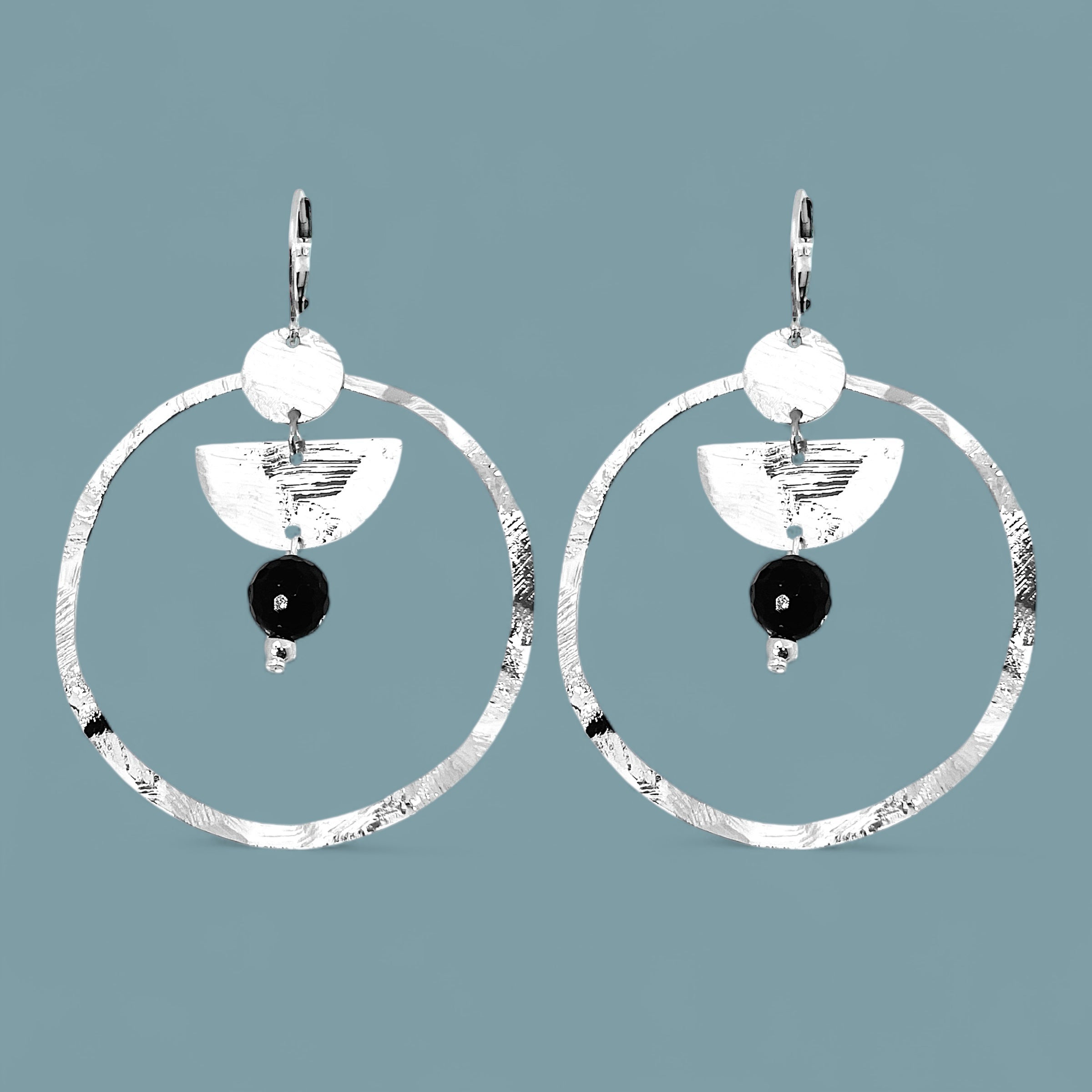 Grandes boucles d'oreilles créoles en métal martelé argenté et pierres naturelles de tourmaline noire accrochée à une demi-lune, sur fond bleu gris