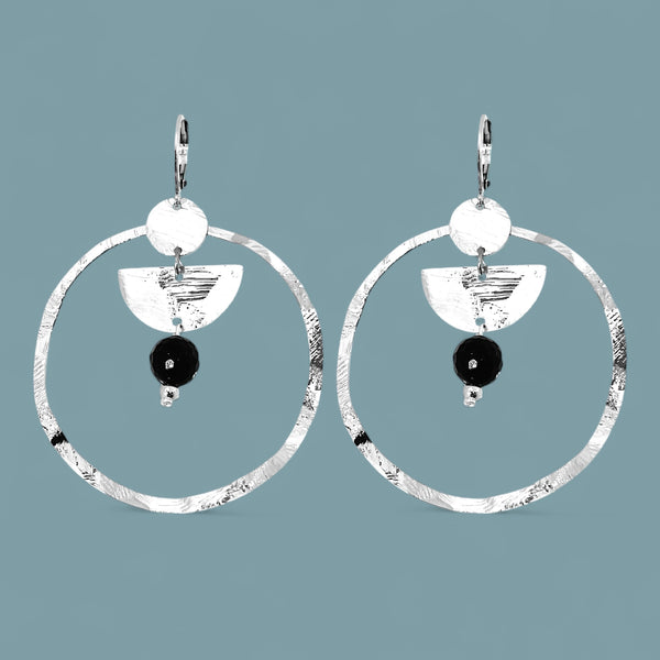 Grandes boucles d'oreilles créoles en métal martelé argenté et pierres naturelles de tourmaline noire accrochée à une demi-lune, sur fond bleu gris