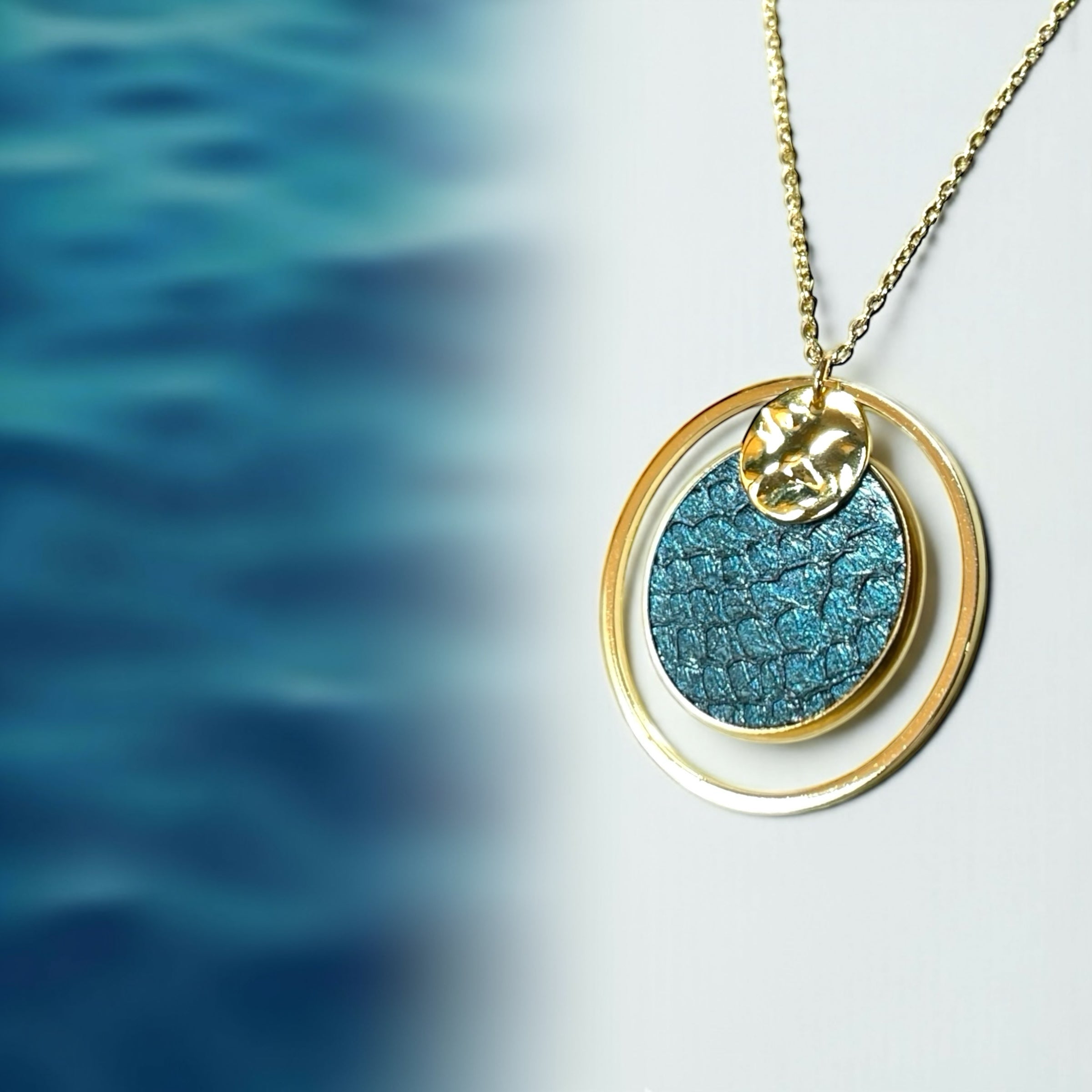 Boucles d’oreilles dorées pendantes Arielles avec pendentif rond en cuir marin bleu paon , sur fond blanc et mer bleu