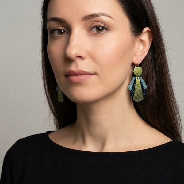 Grandes boucles d'oreilles dorées au design art déco, en cuirs bleu gris nacré, noir à pois holographiques et kaki métallisé, portées aux oreilles par une femme brune