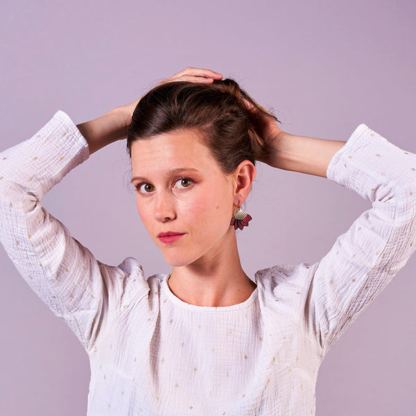 Jeune femme vue de loin portant les boucles d'oreilles dorées en forme éventail en cuir recyclé grenat métallisé modèle Sakura, sur fond mauve