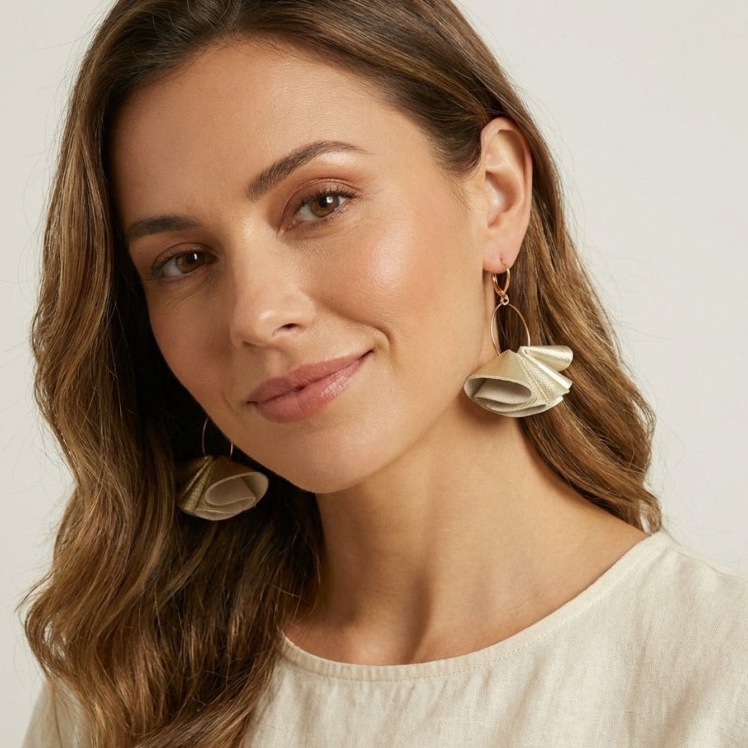Grandes boucles d'oreilles dorées aériennes et sculpturales en cuir plissé doré clair perlé, portées aux oreilles par une femme brune