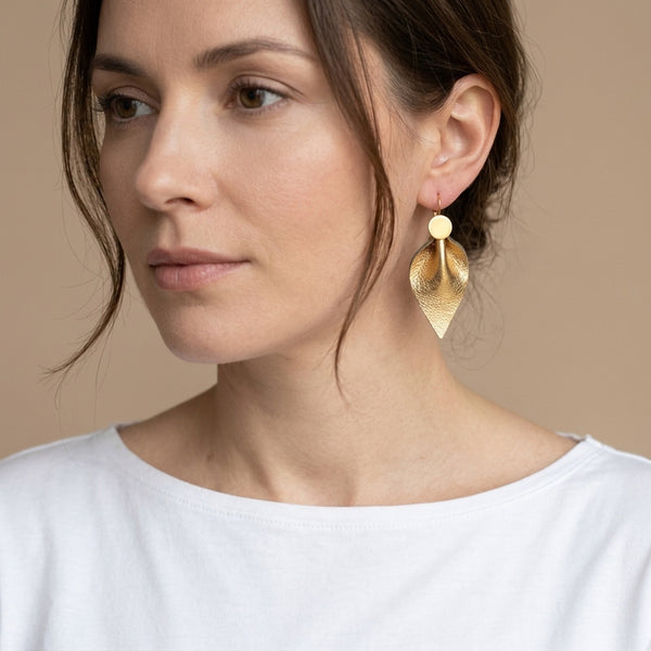 Grandes boucles d’oreilles dorées pendantes en cuir doré antique, en forme de feuille, avec un volume généreux et un effet plissé, portées aux oreilles par ne femme brune