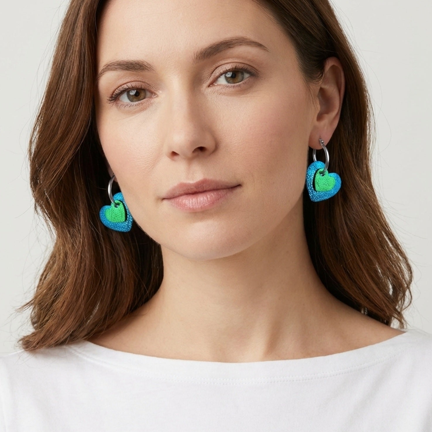 Boucles créoles argentées avec pendentifs en forme de cœurs bleu roi métallisé et vert métallisé, portées à l'oreille par une femme brune