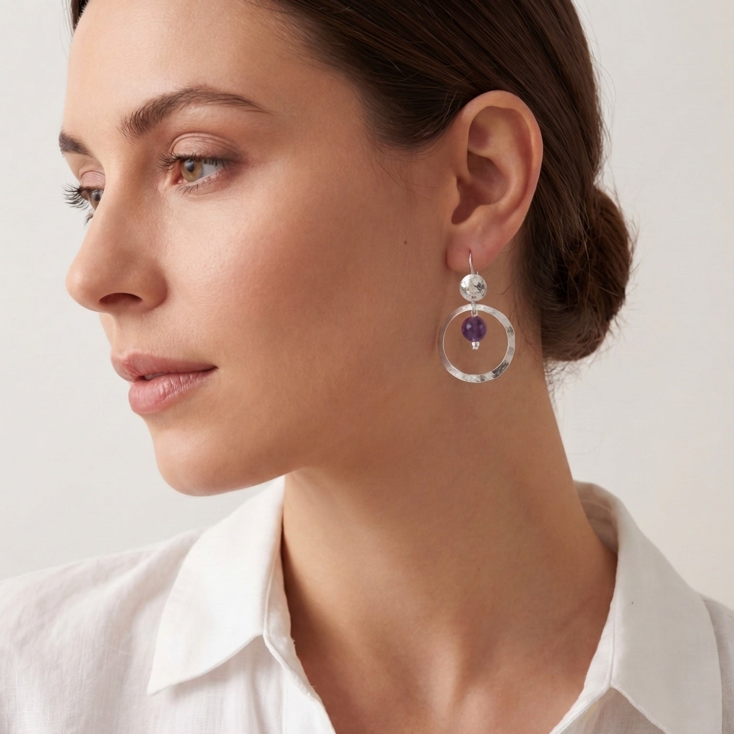 Petites boucles d'oreilles créoles argentées pendantes rondes avec perles d'améthyste, portées à l'oreille par une femme brune