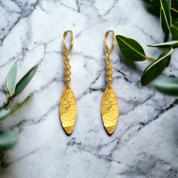 Boucles d'oreilles dorées fines pendantes en pointes, sur marbre gris avec feuilles d'olivier