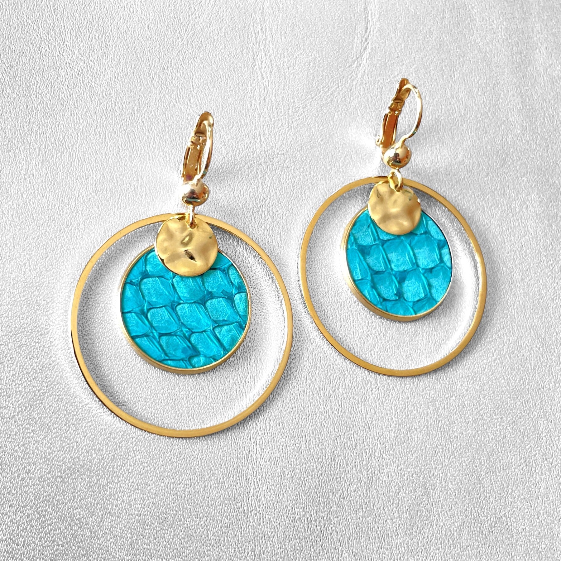 Paire de boucles d'oreilles dorées rondes pendantes de style créoles avec sequin en cuir marin bleu turquoise brut, vue de côté, sur fond gris
