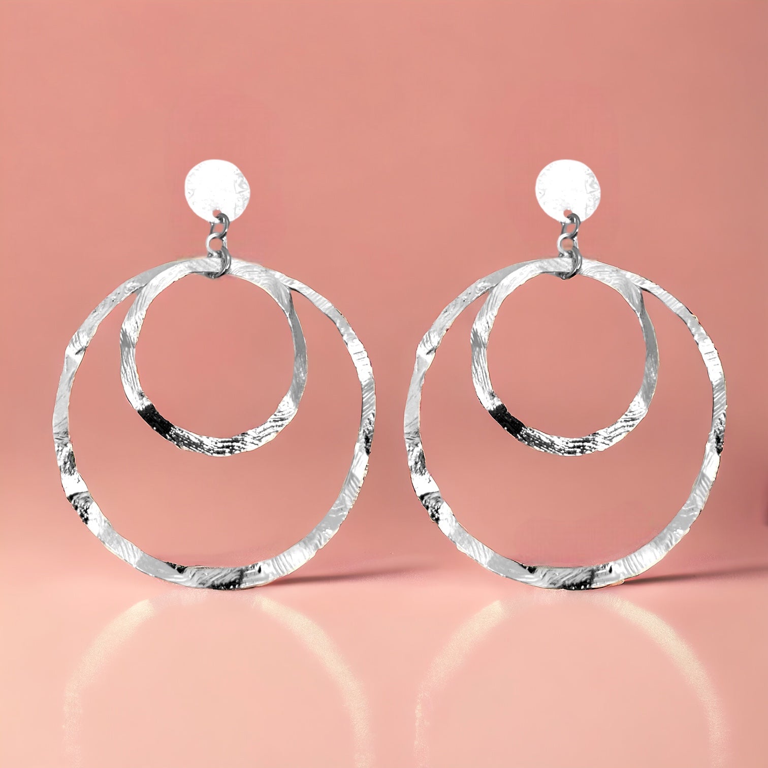 Grandes boucles d'oreilles créoles argentées à double anneau en métal martelé, montées sur puces, sur fond rosé