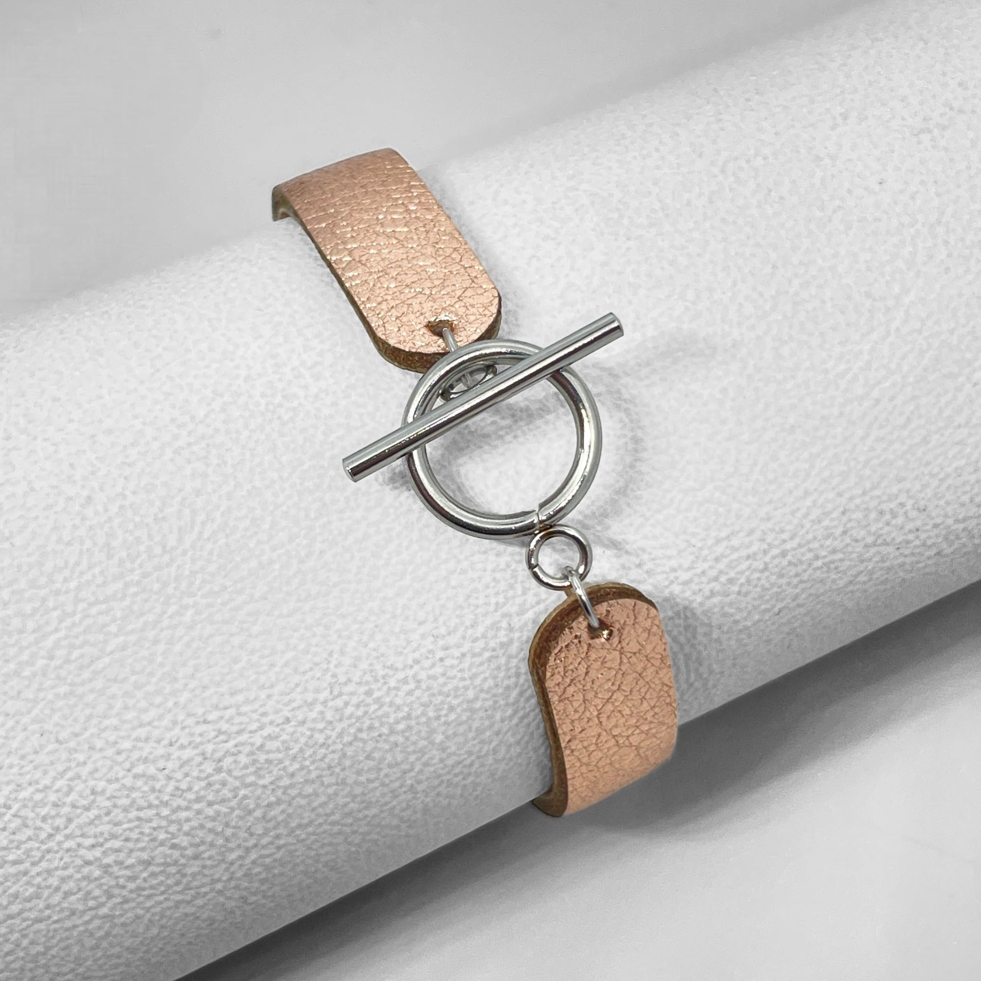 Bracelet en cuir rose pâle métallisé  vu de dessous avec fermoir en T argentée, enroulé autour d'un rouleau de cuir blanc