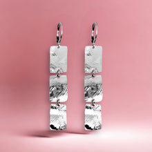 Charger l&#39;image dans la galerie, Boucles d&#39;oreilles pendantes en chapelet de 3 rectangles en métal martelé argenté, sur fond rose
