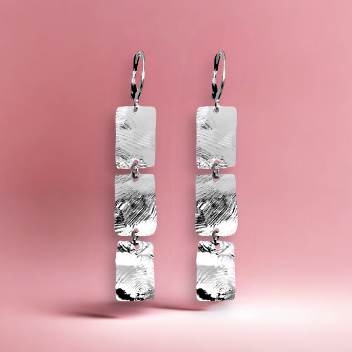 Boucles d'oreilles pendantes en chapelet de 3 rectangles en métal martelé argenté, sur fond rose