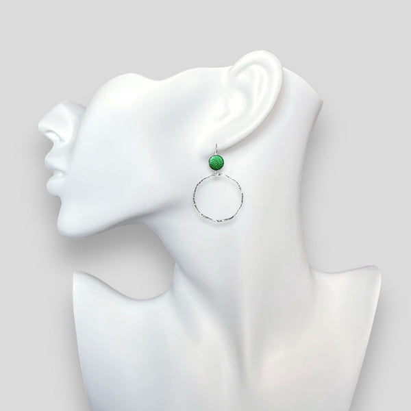 Créoles délicates martelées argentées avec pastille de cuir vert métallisé, portées à l'oreille sur un buste blanc