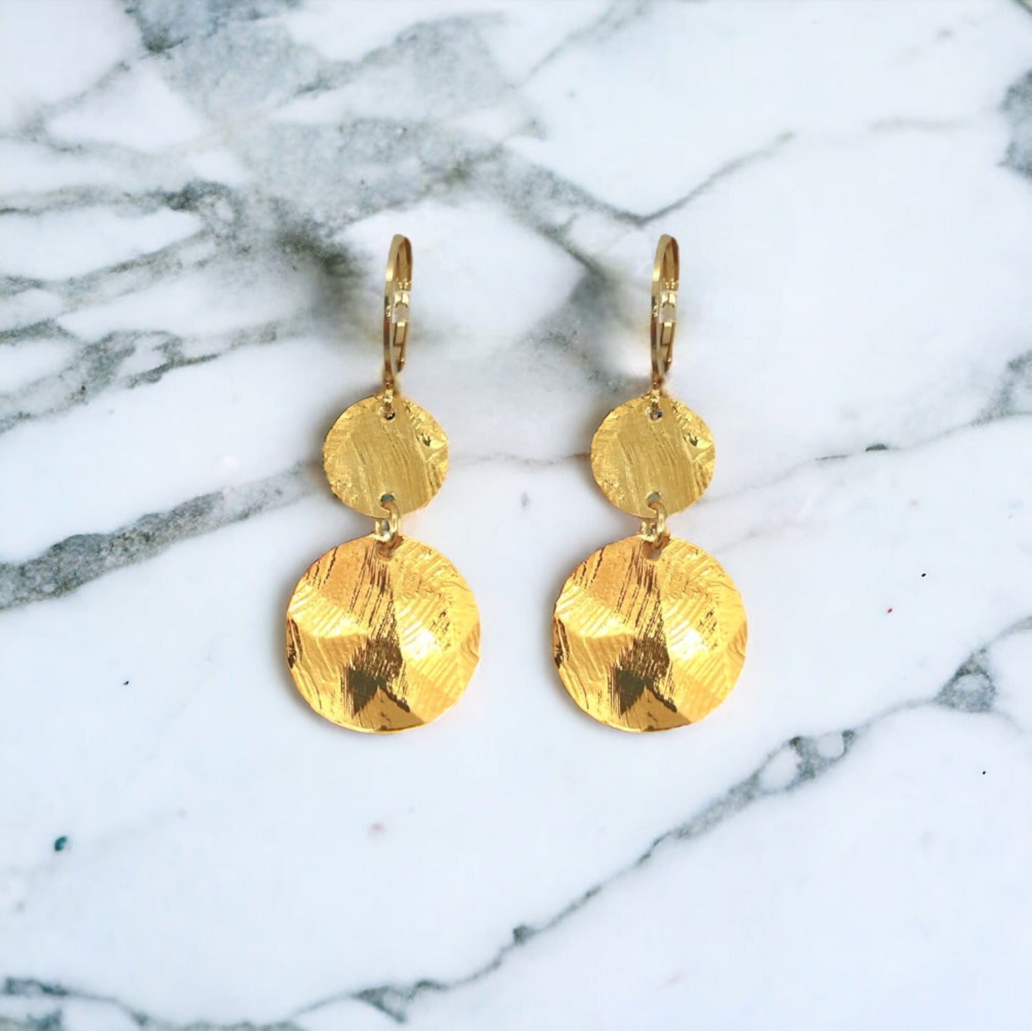 Petites boucles d'oreilles dorées pendantes avec 2 petits sequins ronds en métal martelé, sur fond marbre blanc