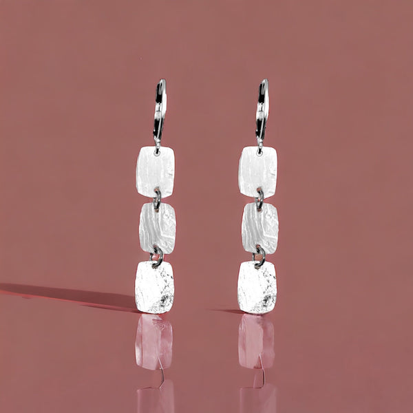 Fines boucles d'oreilles argentées pendantes en chapelet de 3 petits rectangles, sur fond marron clair