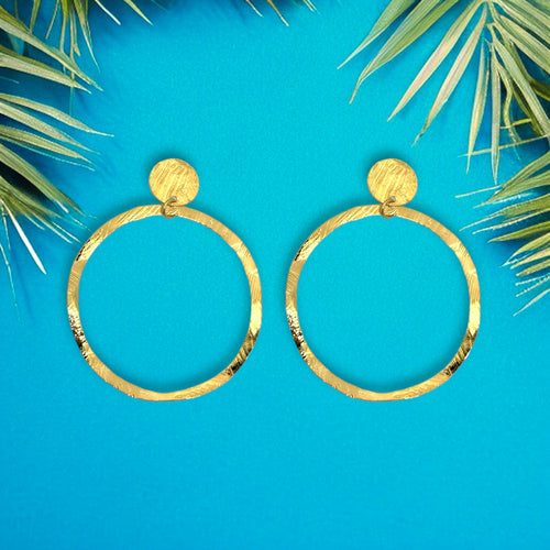 Grandes boucles d'oreilles créoles dorées en métal martelé, montées sur puces rondes, sur fond bleu et feuilles de palmier