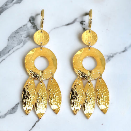 Longues boucles d'oreilles dorées à l'or fin en métal martelé, de style ethnique bohème chic, avec leurs pampilles en forme de plumes, sur marbre blanc