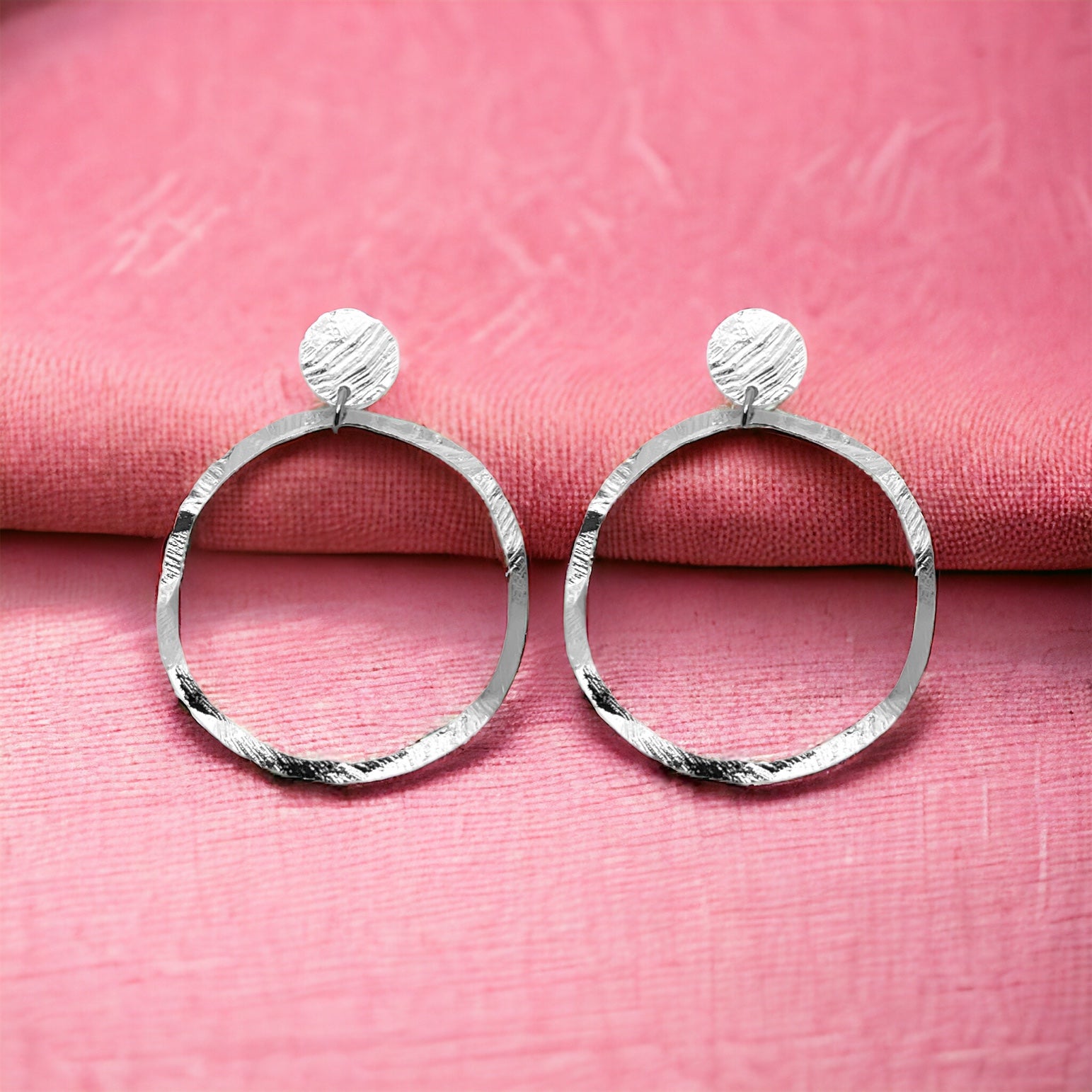 Grandes boucles d'oreilles créoles argentées en métal martelé, montées sur puces rondes, sur tissu rose