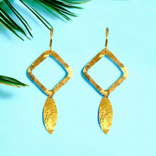 Charger l&#39;image dans la galerie, Boucles d&#39;oreilles dorées avec pendentif en forme de losanges et pointes, sur fond bleu et feuilles de palmier
